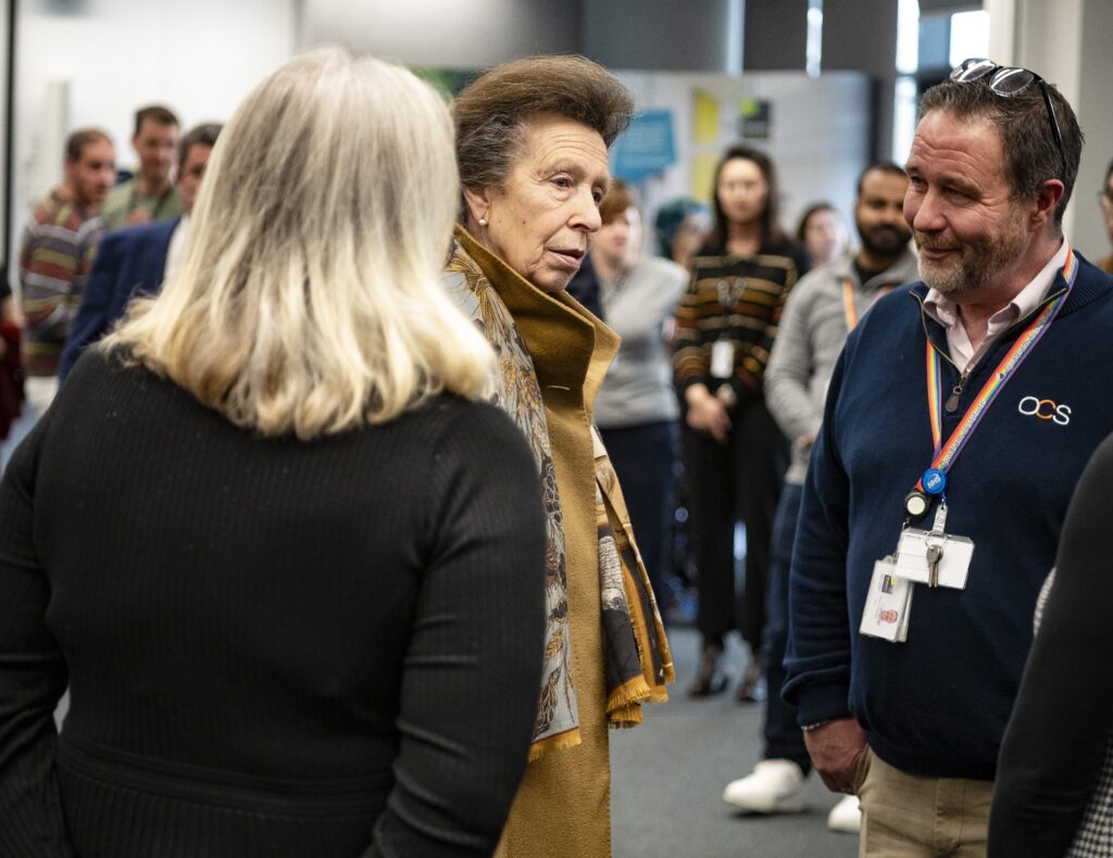 A woman in a brown coat and scarf speaks with two people in an office space, while others stand in the background, watching the interaction.