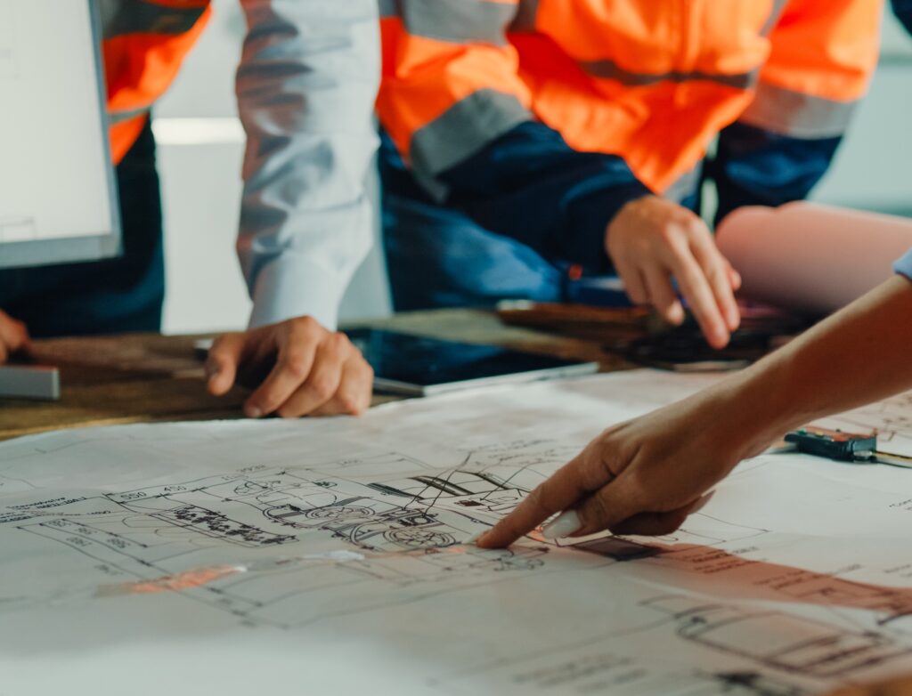 Close-up of people wearing orange safety vests reviewing technical drawings or blueprints spread out on a table, with one person pointing to a specific detail on the paper.