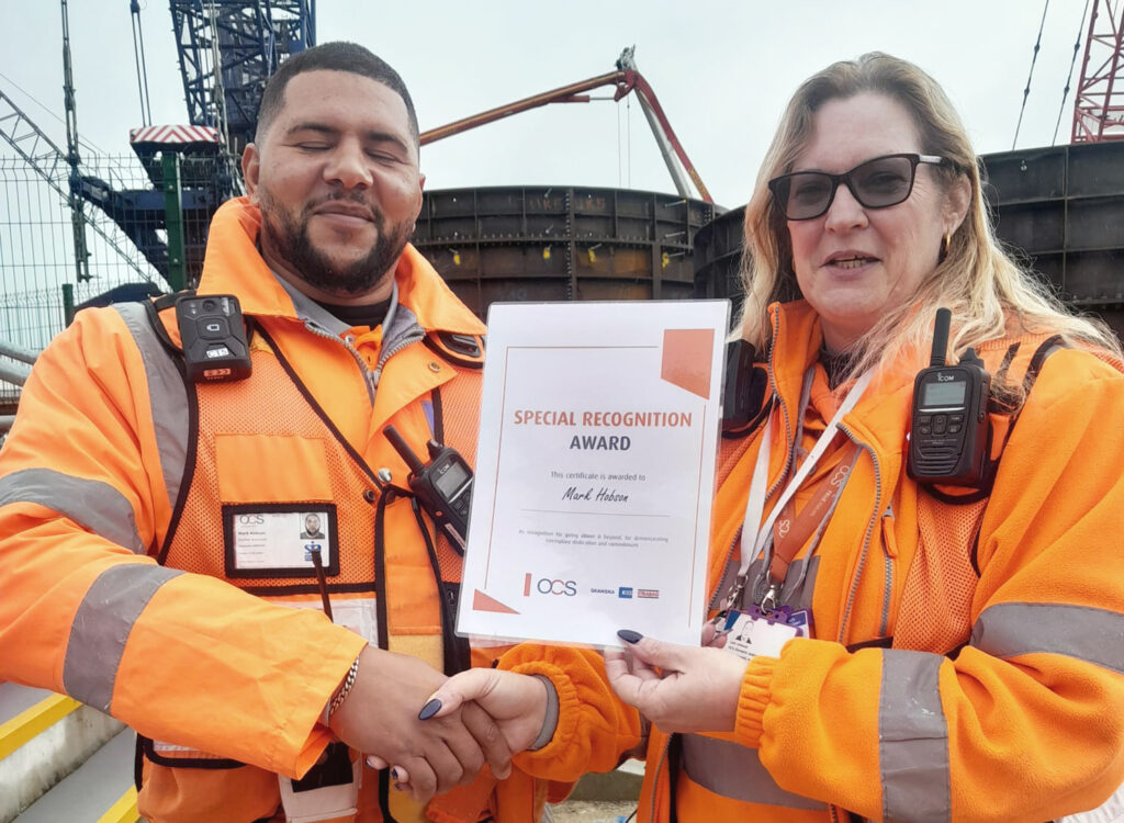 Two people in orange safety jackets and radios shake hands at a construction site. One person holds a Special Recognition Award certificate. Both are smiling, with industrial equipment visible in the background.