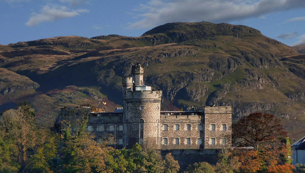 A stone castle with square towers sits among autumn trees, backed by rolling, rugged hills under a partly cloudy sky.