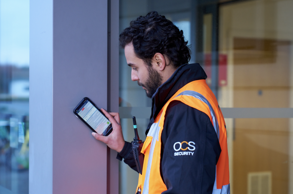 A security guard in an orange OCS Security jacket stands beside a building, looking at a smartphone screen. He holds the phone in his left hand and has a radio clipped to his jacket.