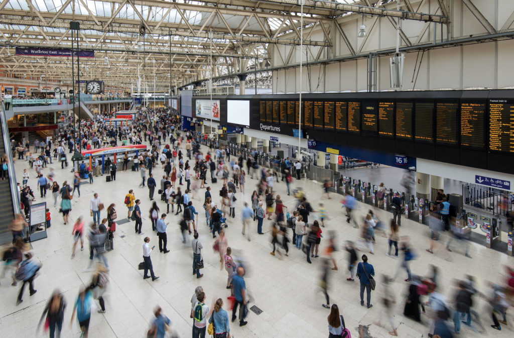 A busy train station concourse with crowds of people walking in various directions. Large electronic departure boards display train schedules above ticket counters. The station has a high glass roof and metal beams.