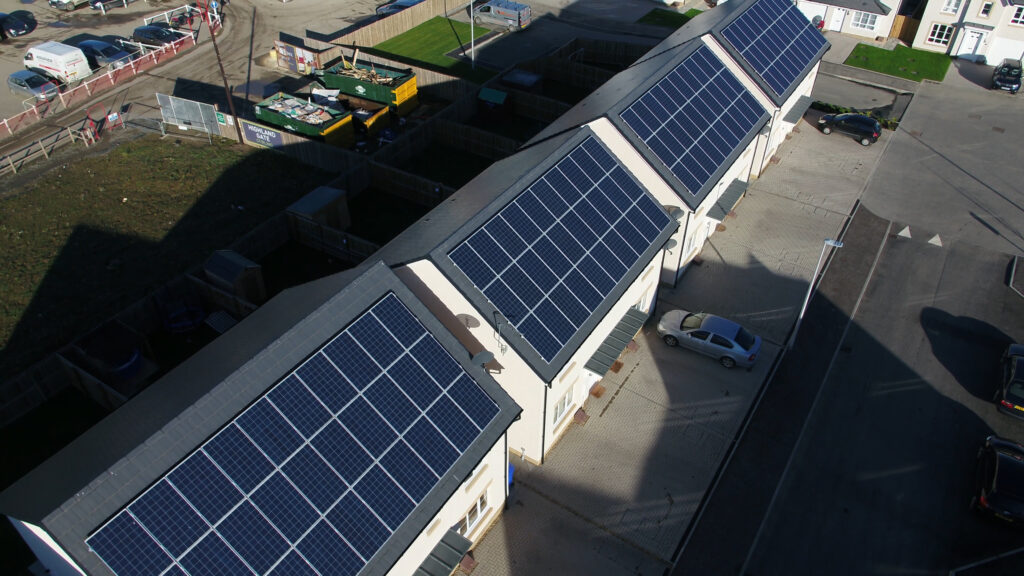 A row of houses with pitched roofs covered in solar panels, seen from above in daylight. A white car is parked beside one house, and nearby areas include some grass, fencing, and industrial containers.