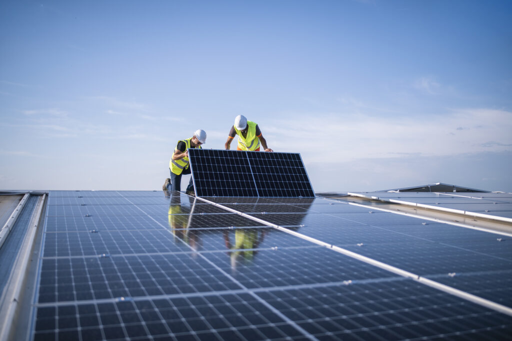 Two workers in safety vests and helmets install a solar panel on a rooftop under a clear blue sky, surrounded by other solar panels.