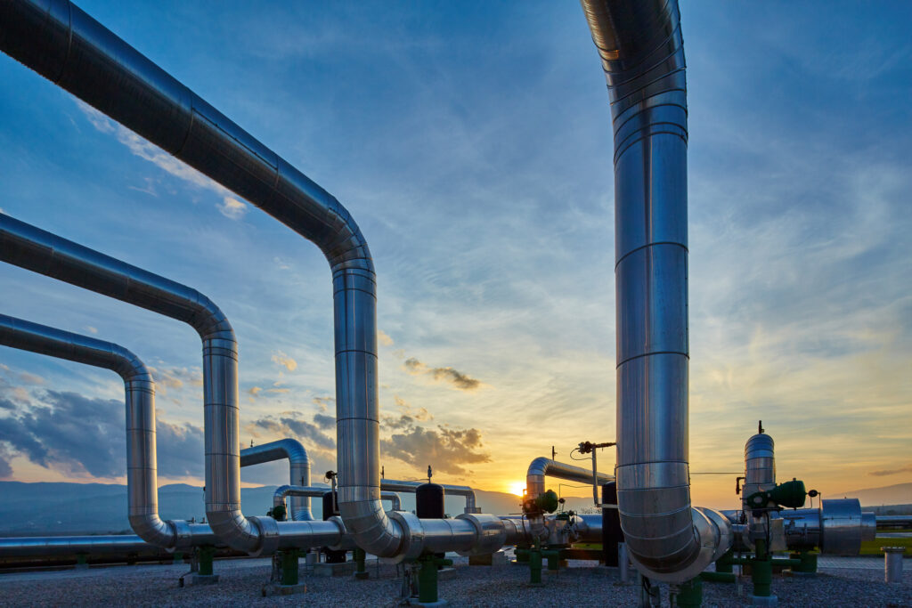 Large metallic industrial pipelines stretching across an outdoor facility, silhouetted against a sunset sky with scattered clouds, and mountains visible in the background.
