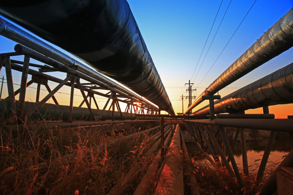 A low-angle view of large industrial pipelines elevated on metal supports, with the sunset sky in the background and tall grasses in the foreground. Power lines run parallel to the pipes.