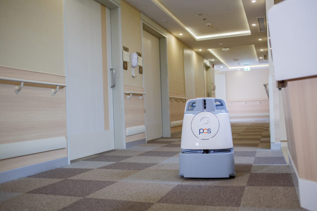 A small, white delivery robot with the logo PQS is positioned in the hallway of a modern building with beige walls and doors, possibly a hospital or care facility.