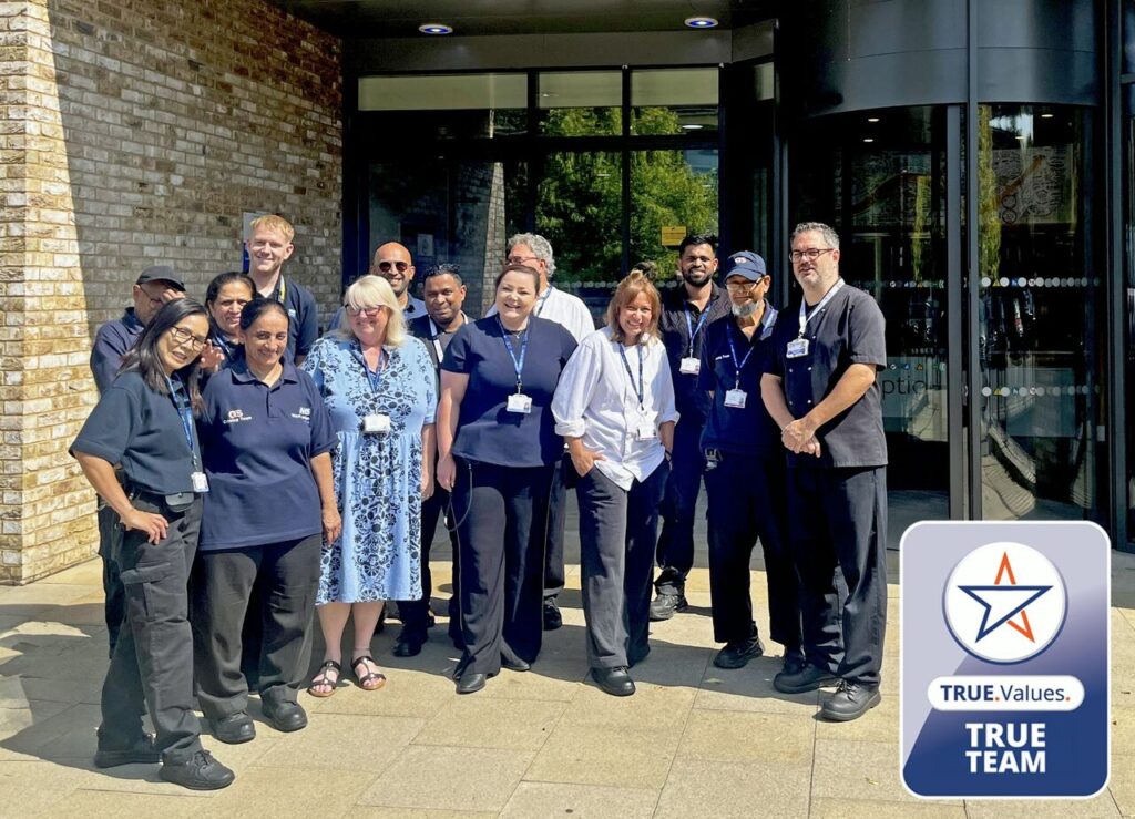 A diverse group of people in work uniforms and badges stand smiling outside a building. One person wears a blue dress. At the bottom right, there is a badge logo that reads “TRUE Values. TRUE TEAM.”.