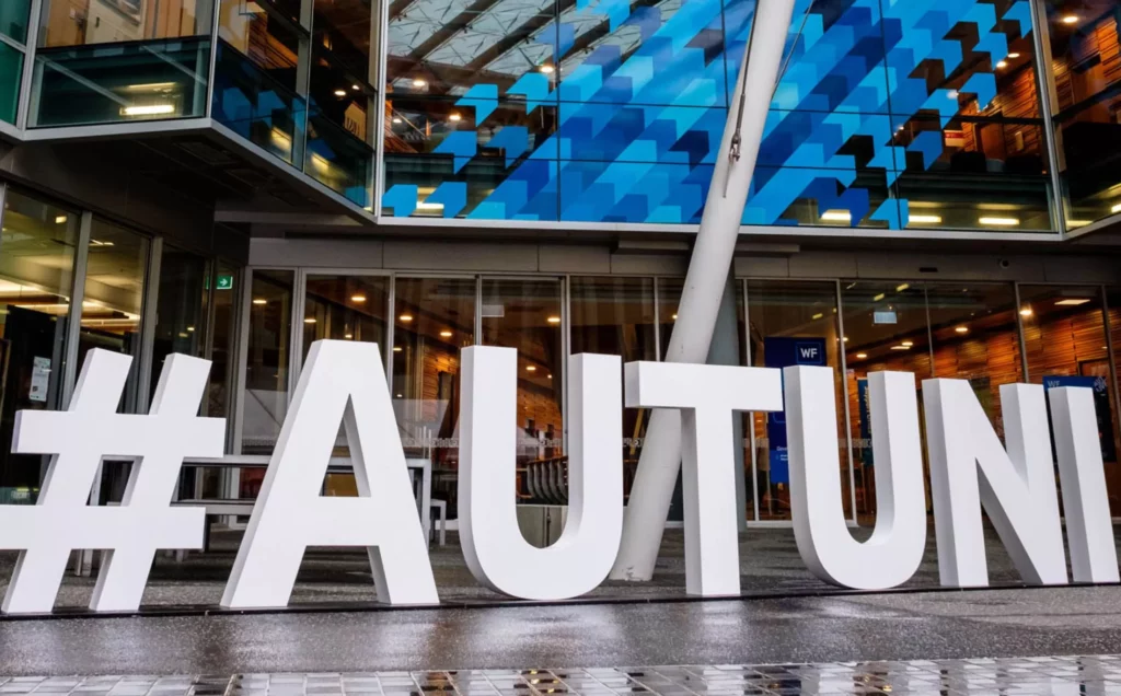Large white hashtag letters spell out #AUTUNI in front of a modern glass building with blue geometric patterns and reflections, on a wet paved surface.