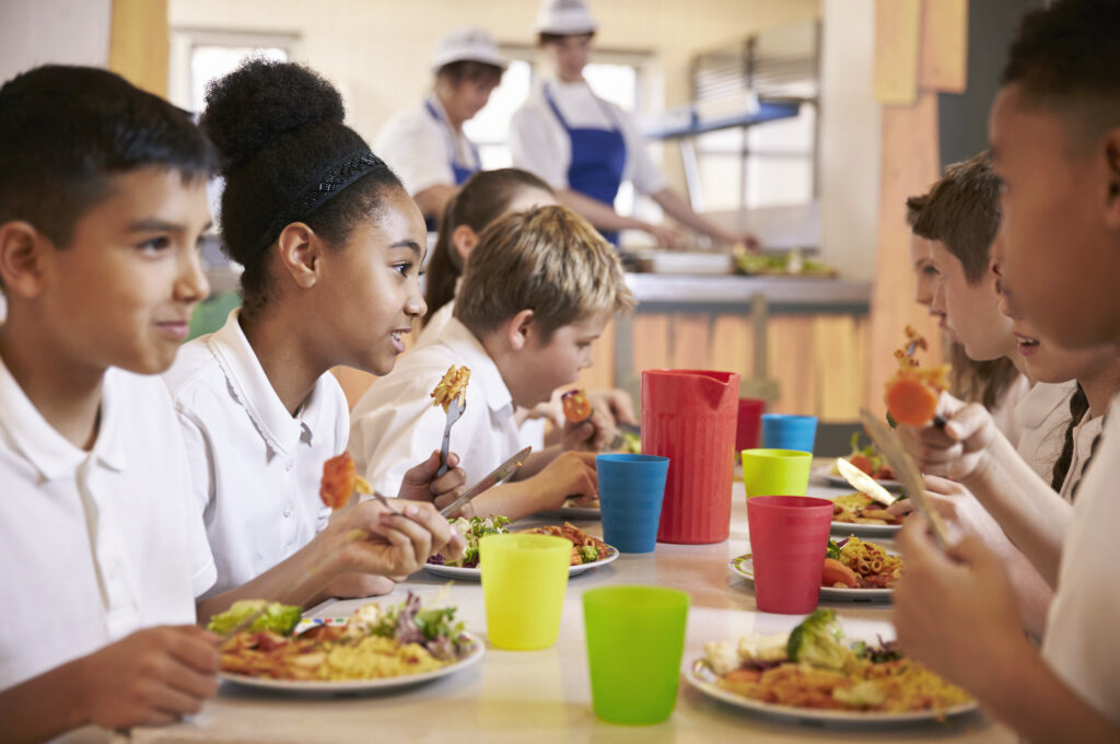 A group of schoolchildren sit together at a cafeteria table, eating lunch and talking. Colorful cups and plates of food are in front of them, and cafeteria staff are visible in the background.