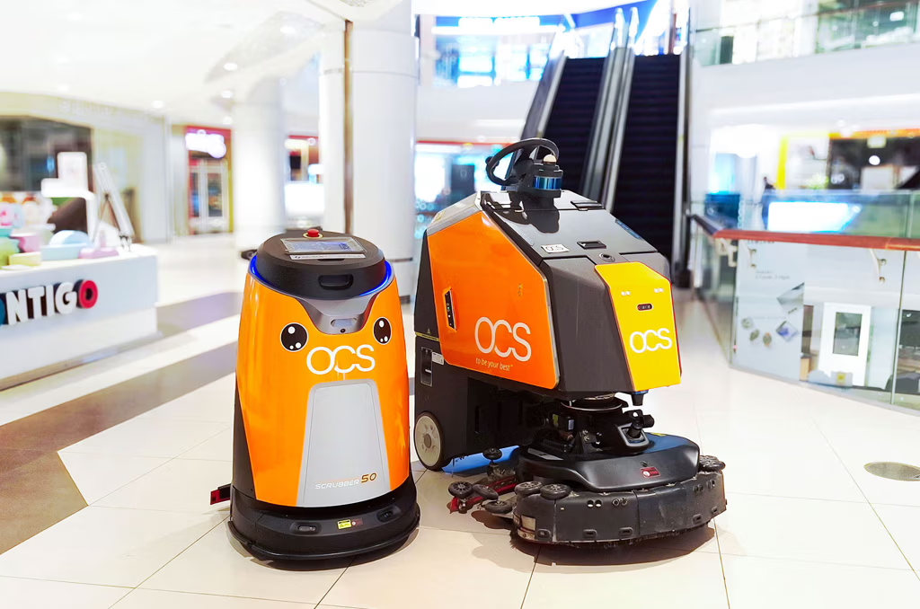 Two orange and black floor-cleaning robots with OCS branding stand side by side in a shopping mall near an escalator. One robot has a cute face design, while the other has a seat for a human operator.