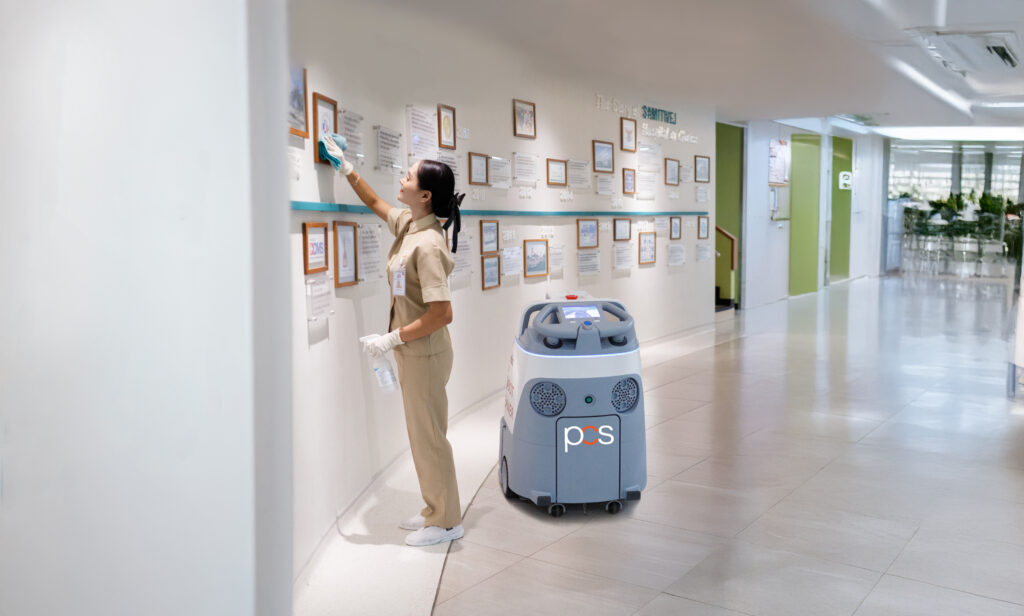 A woman in a beige uniform cleans framed documents on a wall in a bright, modern hallway, with a large cleaning machine labeled pcs beside her.