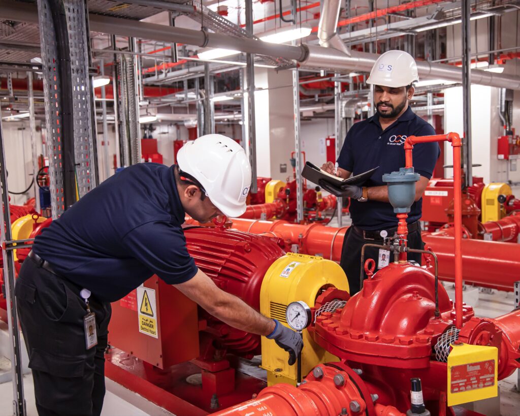 Two workers in hard hats inspect and take notes on red industrial equipment with pipes and gauges in a facility. One checks a pressure gauge while the other writes on a clipboard. Safety signage is visible.