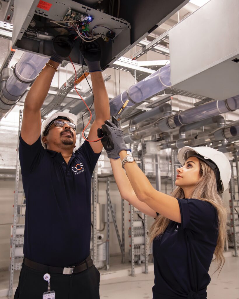 Two engineers in safety helmets and gloves work together on electrical wiring in an industrial setting. One adjusts wires overhead while the other assists, both wearing navy shirts and safety glasses in a modern facility.