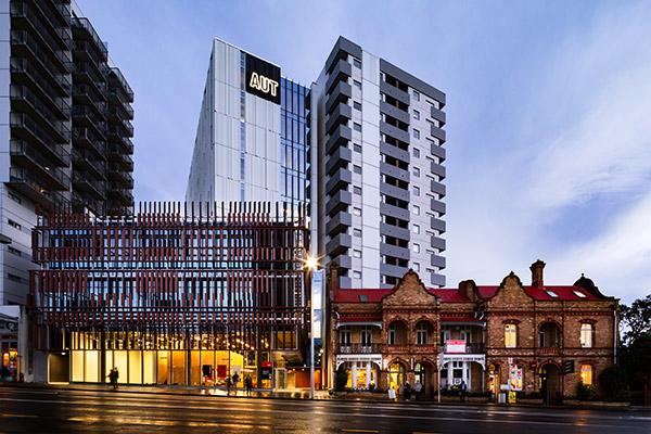 A modern glass building with AUT signage stands beside tall apartment blocks and a historic brick building with red roofs, all facing a wet street at dusk.