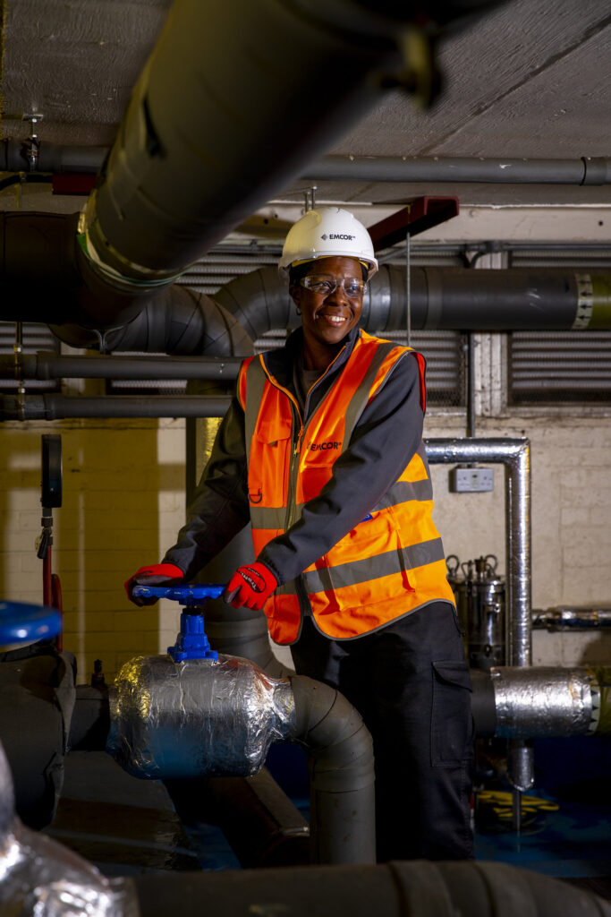 A smiling worker wearing a white hard hat, orange safety vest, and red gloves operates a large blue valve on industrial pipes in a dimly lit mechanical room.