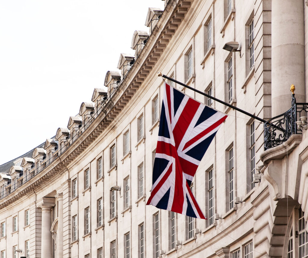A Union Jack flag hangs from a pole on the exterior of a curved, white stone building with many windows and classical architectural details.