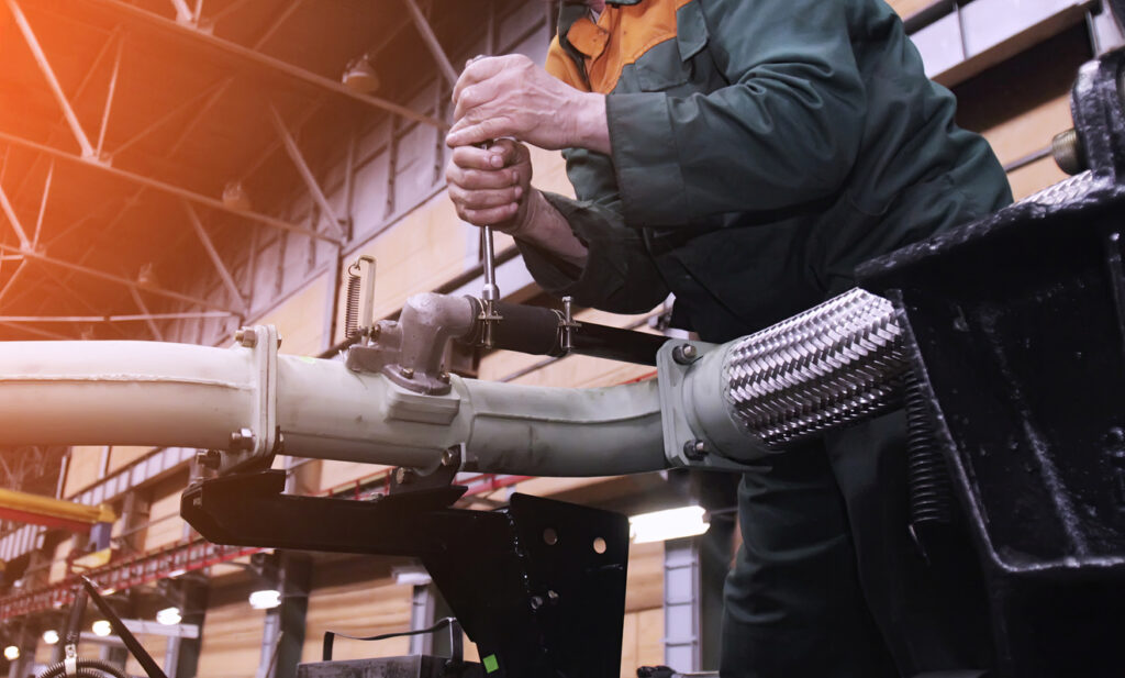 A worker in green overalls uses a wrench to tighten a metal pipe fitting inside a large industrial facility with bright overhead lights.