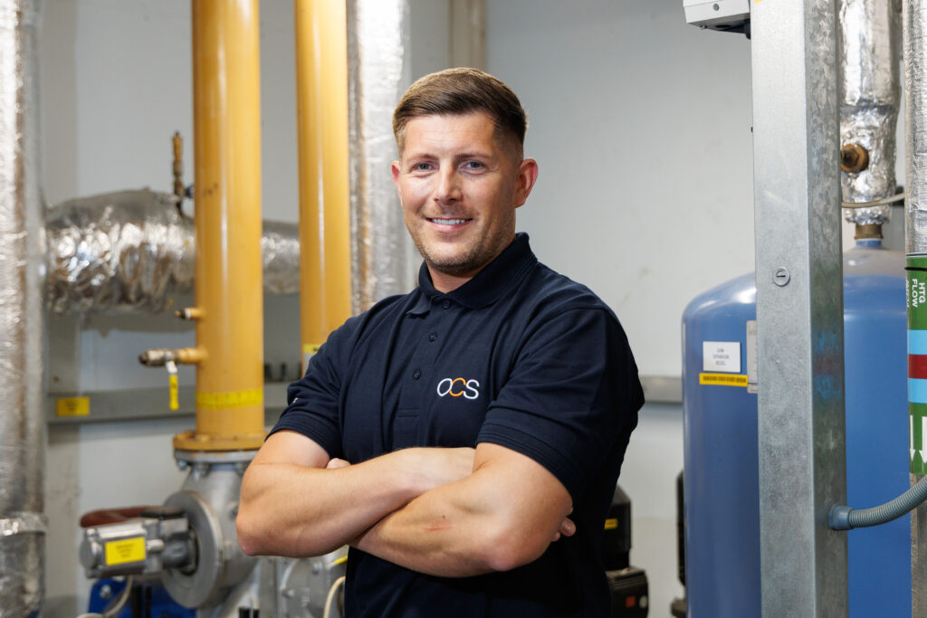 A man in a navy blue polo shirt stands with arms crossed, smiling, in front of industrial pipes and machinery in a utility or boiler room.