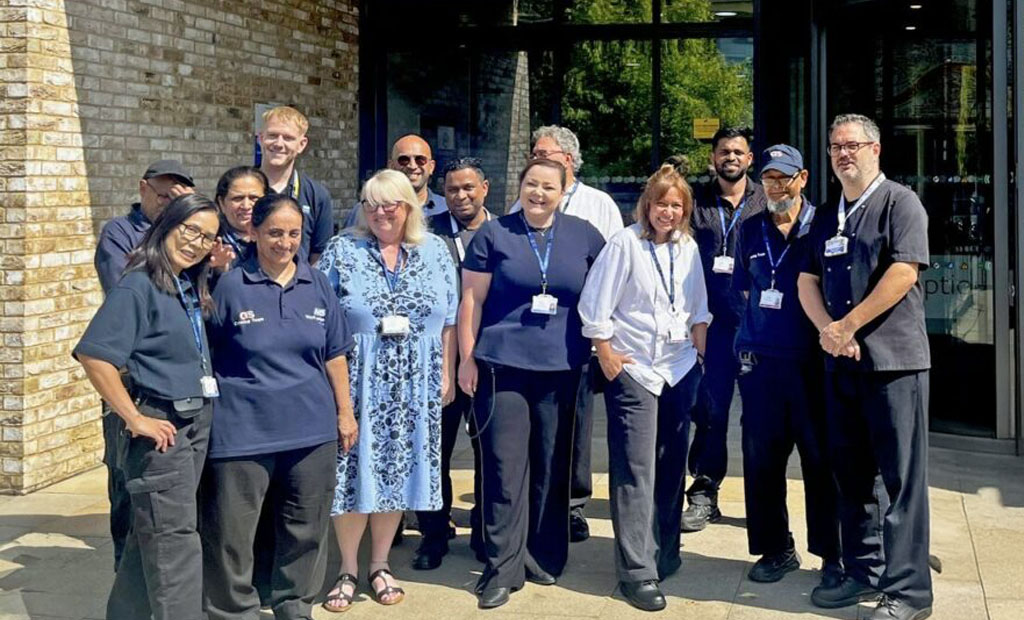 A group of thirteen people, some in uniforms and others in casual clothing, stand smiling together outside a building with glass doors and a brick wall on a sunny day.