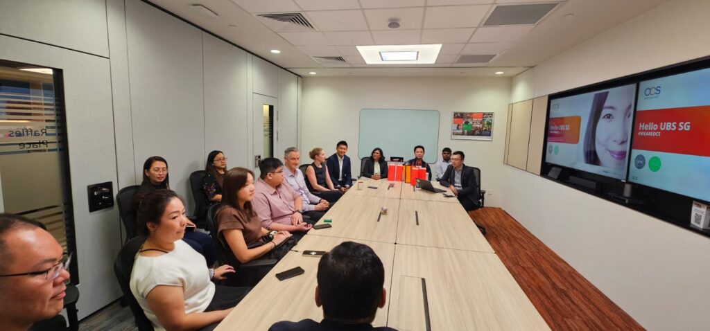 A group of people in business attire sit around a long conference table in a modern office meeting room, with a large screen displaying Hello UBS SG at the front.