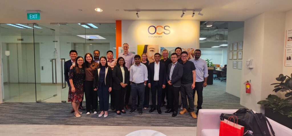 A group of sixteen people, both men and women, stand smiling in an office lobby with glass doors and an OCS sign on the wall behind them. Some plants and office furniture are visible in the foreground.