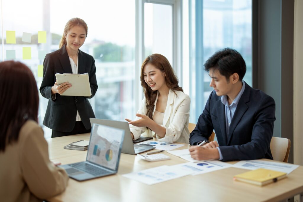 Four professionals in business attire sit around a conference table with laptops and documents. One woman stands holding a clipboard, while the others engage in discussion, appearing collaborative and focused.