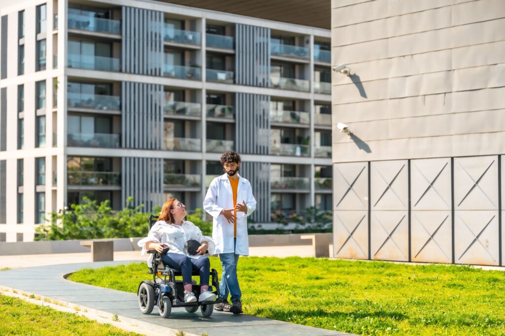 A woman in a wheelchair smiles and talks with a man in a white coat as they travel along a path outside a modern building with balconies. They appear to be enjoying a pleasant day.