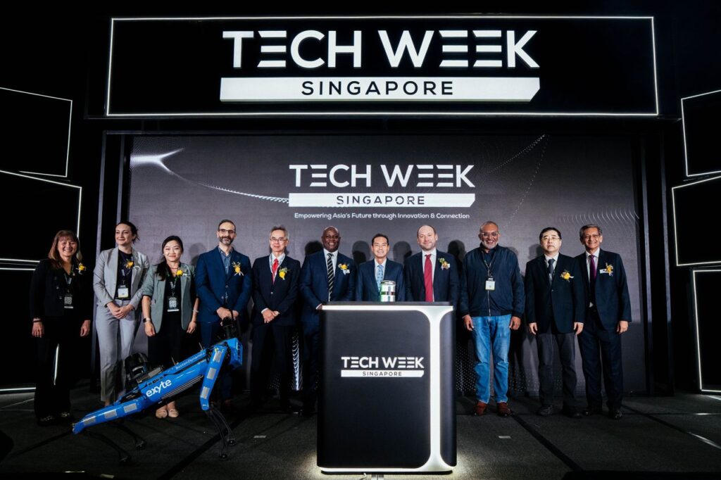 A group of people in business attire stand on stage under a Tech Week Singapore sign, with a robotic dog nearby and a podium in front, at a technology conference event.