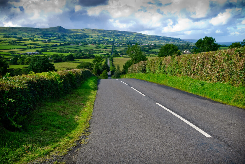 A narrow rural road lined with green hedges stretches into the distance, leading to rolling hills under a partly cloudy sky in a lush countryside landscape.