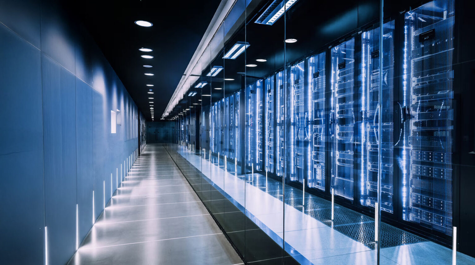 A modern data center hallway with glass walls on one side, revealing rows of illuminated server racks and computer equipment glowing with blue lights. The space is clean, high-tech, and well-lit.