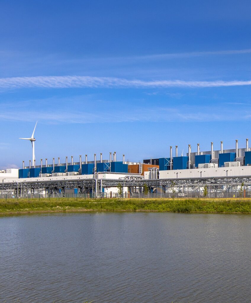 A modern industrial facility with blue and gray buildings and many smokestacks stands behind a body of water, with a wind turbine visible on the left under a clear blue sky.