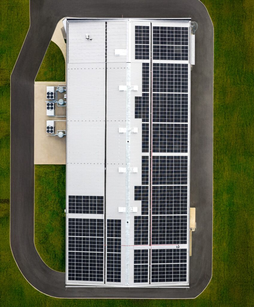 Aerial view of a large rectangular building with a roof covered in solar panels, surrounded by a paved road and green grass. HVAC units are visible on a platform next to the building.