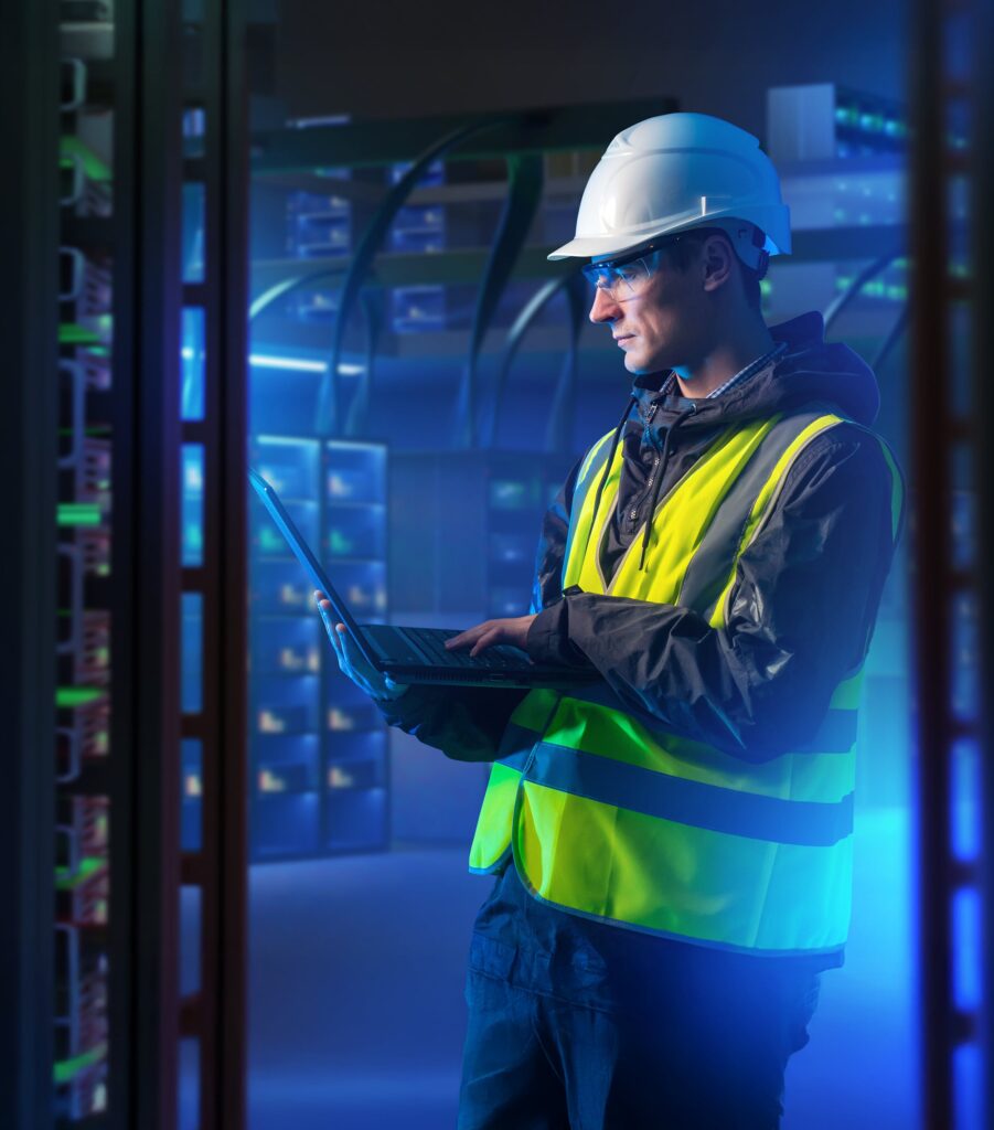 A worker wearing a hard hat, safety glasses, and a reflective vest uses a laptop in a server room filled with glowing computer racks and blue lighting.