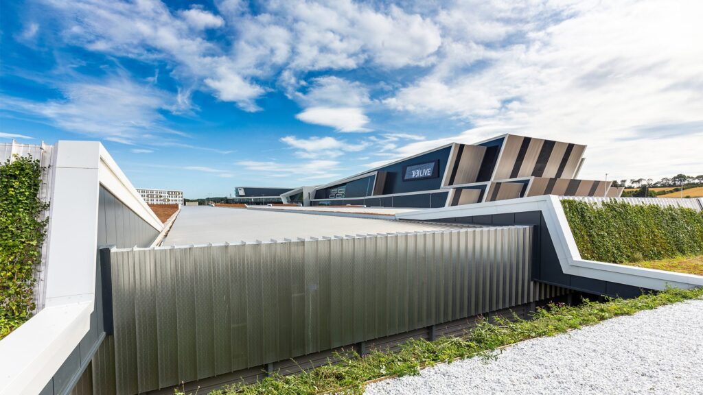 A modern building with geometric architecture, angled walls, and metallic panels, set against a bright blue sky with scattered clouds. Greenery lines the edges of the structure.