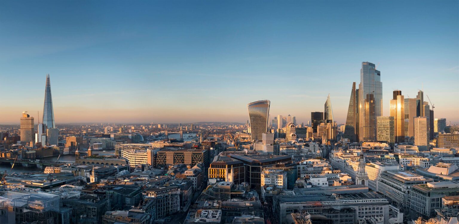A panoramic view of London’s skyline at sunset, featuring modern skyscrapers like The Shard and the Walkie Talkie building, with the River Thames and historic city buildings visible under a clear blue sky.
