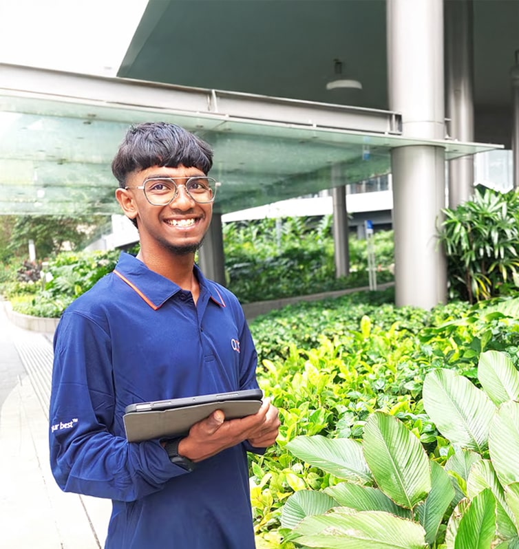 A young man wearing glasses and a blue shirt stands outdoors by lush green plants, smiling and holding a tablet. Modern buildings with glass and metal elements are visible in the background.