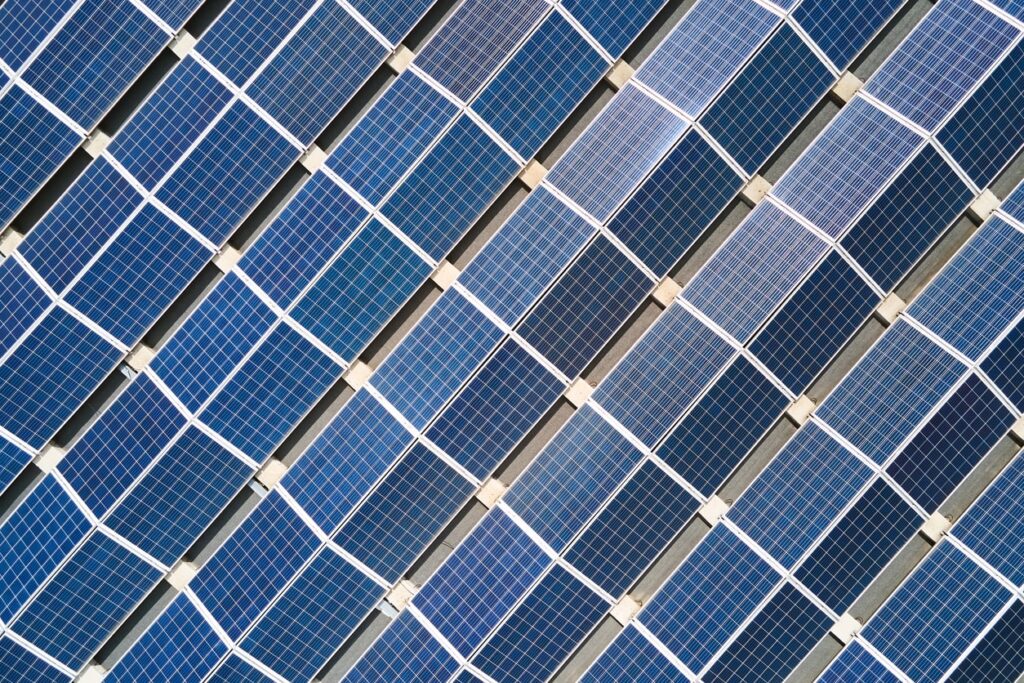 Aerial view of rows of blue solar panels arranged diagonally, forming a geometric pattern. The panels are divided by thin white lines and are installed outdoors, capturing sunlight for renewable energy.