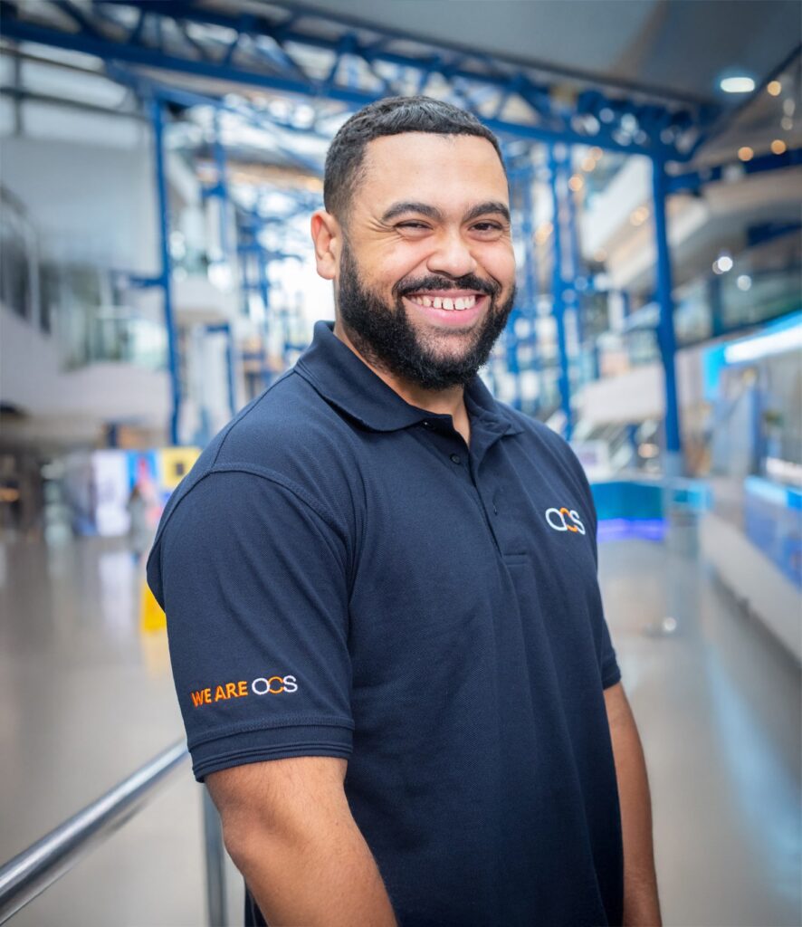 A man with a beard smiles while standing indoors, wearing a navy blue polo shirt with OCS and WE ARE OCS logos. The background features modern architecture with blue beams and glass.