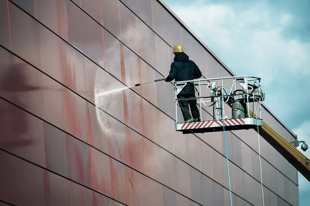 A worker wearing a yellow helmet and safety gear uses a high-pressure hose to clean the exterior of a large red building while standing on a mechanical lift against a cloudy sky.