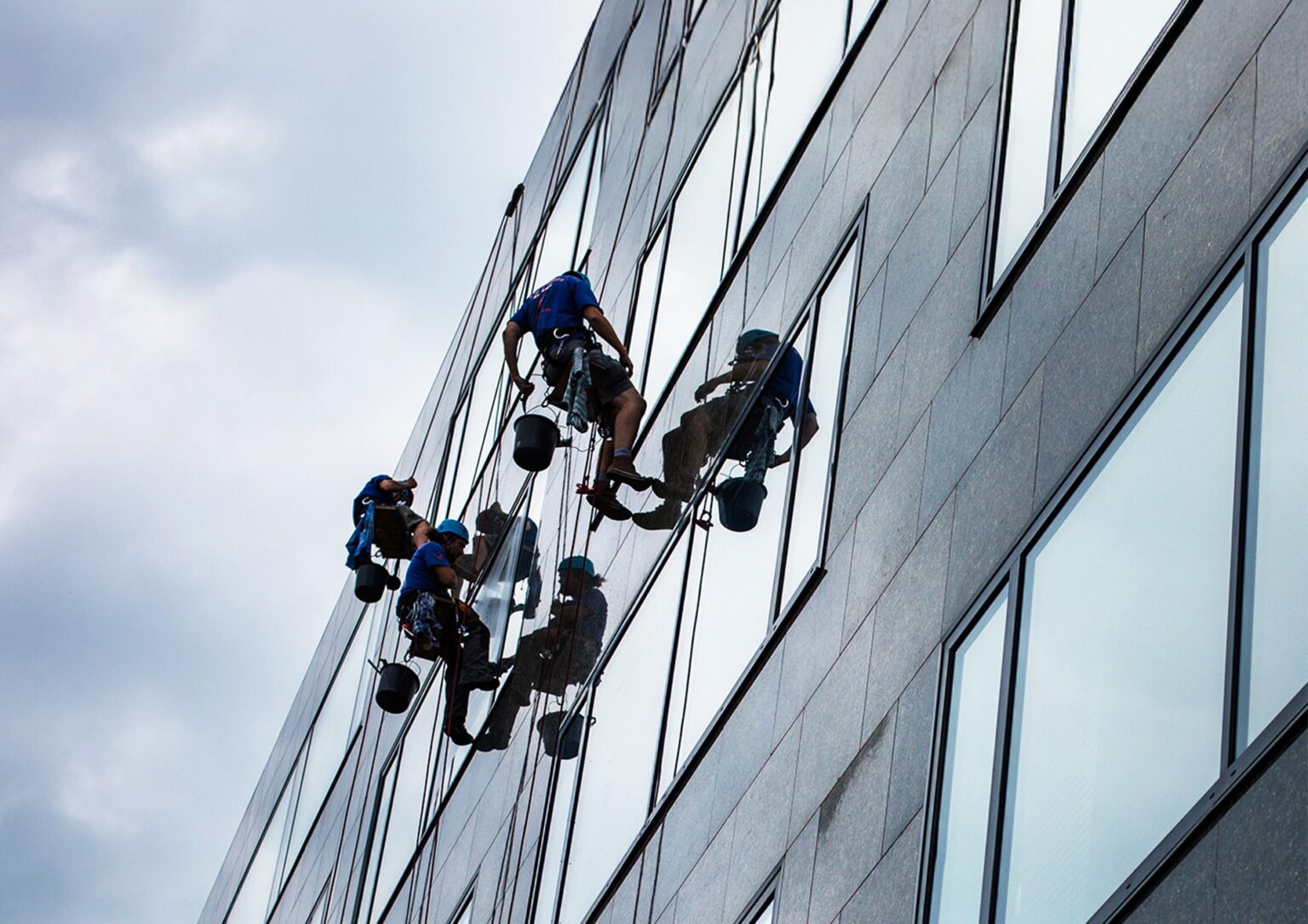Three workers wearing safety harnesses and helmets are suspended on ropes, cleaning the windows of a tall glass building. Reflections of the workers are visible in the windows. The sky is cloudy.