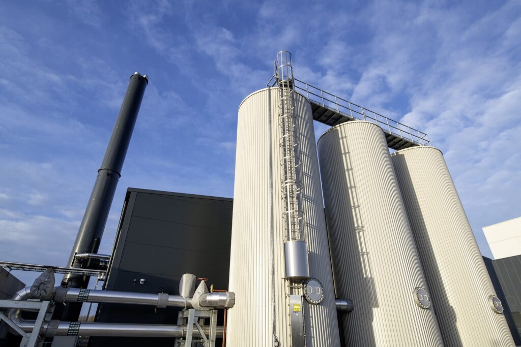 Large cylindrical industrial storage tanks with ladders and pipes attached, set against a blue sky with scattered clouds. A tall black smokestack and a dark building structure are also visible in the background.