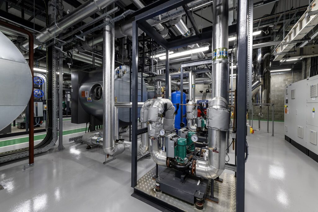 A modern industrial mechanical room with metal pipes, valves, pumps, control panels, and machinery, all neatly arranged on a polished gray floor with visible overhead lighting and wiring.