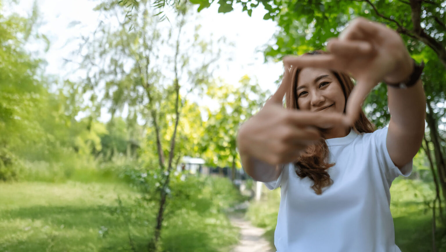 A smiling woman in a white shirt stands outside on a sunny day, holding her hands up to form a rectangular frame. She is surrounded by green trees and grass, with a blurred path in the background.