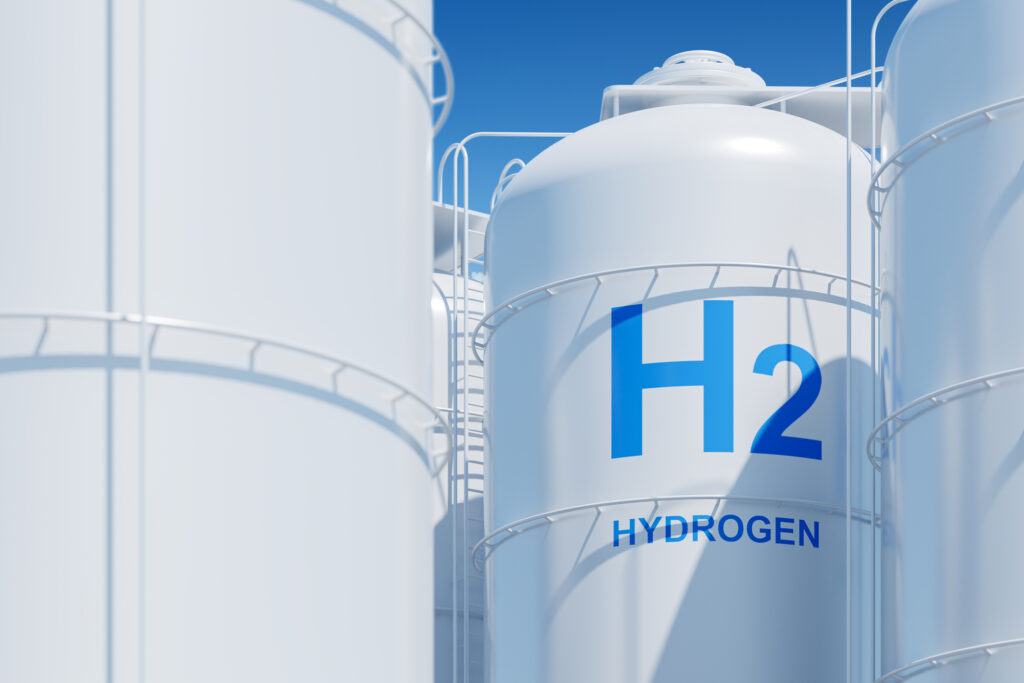 Large white storage tanks with “H2 HYDROGEN” written in blue on one tank, surrounded by metal railings and ladders, under a clear blue sky; representing hydrogen gas storage facilities.