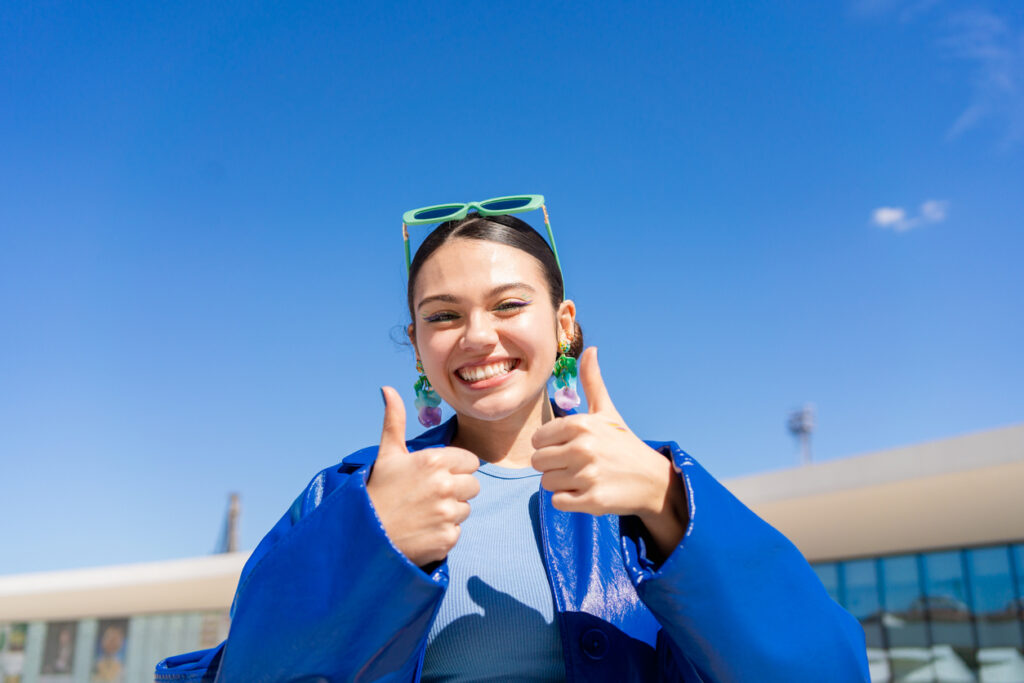 A smiling woman wearing a bright blue jacket and green sunglasses gives two thumbs up outdoors under a clear blue sky.
