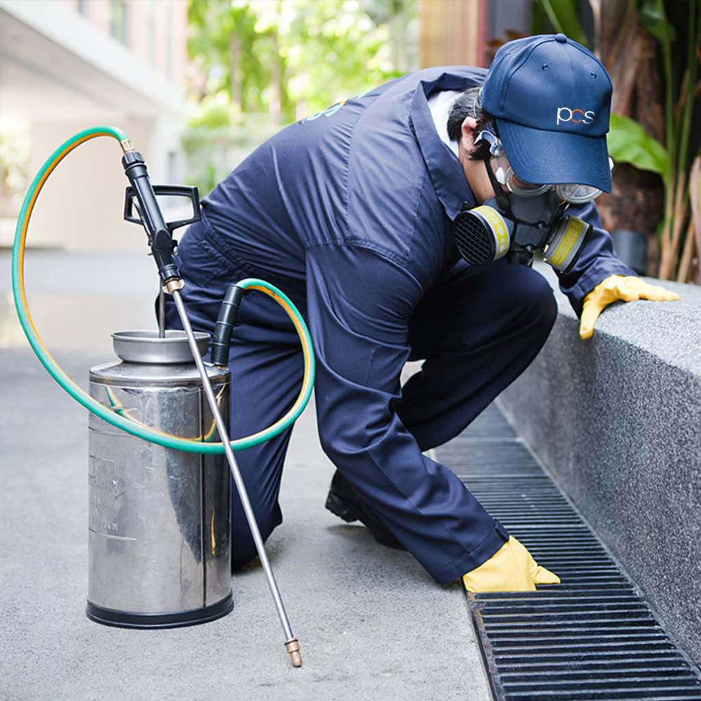 A pest control worker in protective gear kneels by a building’s drain, inspecting it while holding a metal pesticide sprayer. Greenery and building structures are visible in the background.
