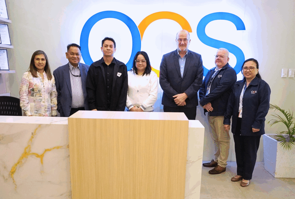 Seven people stand in a row behind a marble reception desk, posing for a group photo in front of a wall with a large OCS logo. Four men and three women are dressed in business attire and business casual clothing.