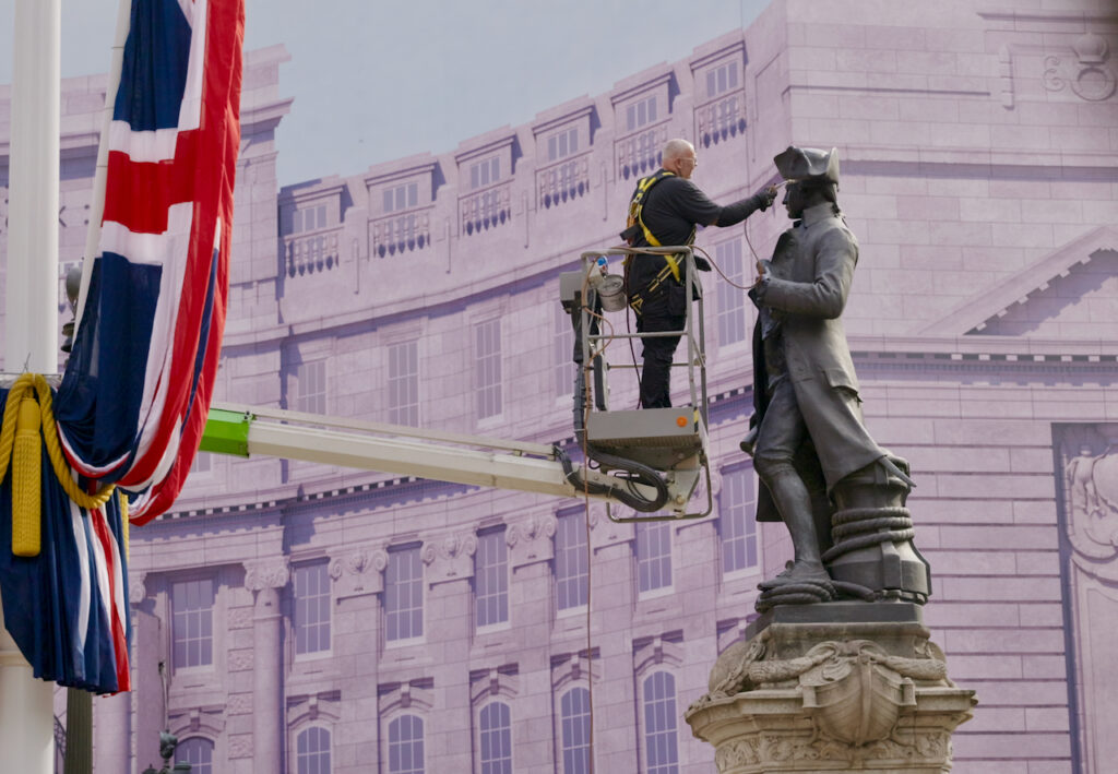A worker on a mechanical lift cleans or repairs a large statue while wearing safety gear. A draped British flag is visible to the left, and an ornate stone building stands in the background.