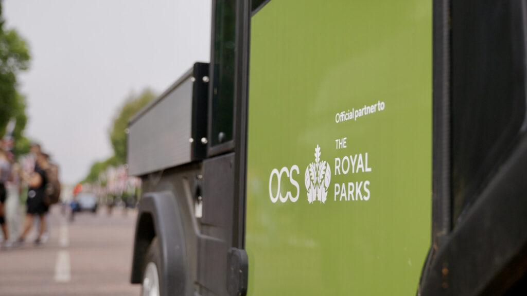 A green utility vehicle with Official partner to OCS The Royal Parks written on the side is parked outdoors, with people walking and trees visible in the blurred background.
